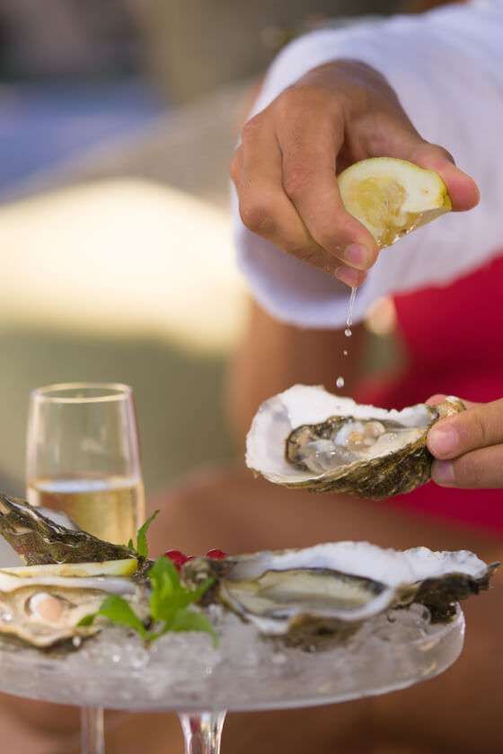 A hand squeezes a lemon wedge over a fresh oyster on a bed of crushed ice, with a glass of white wine in the background.