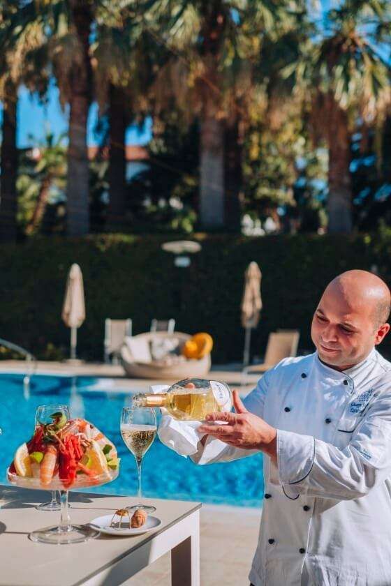 A chef pours wine into a glass next to a seafood platter on a table by a sunny pool with palm trees in the background.