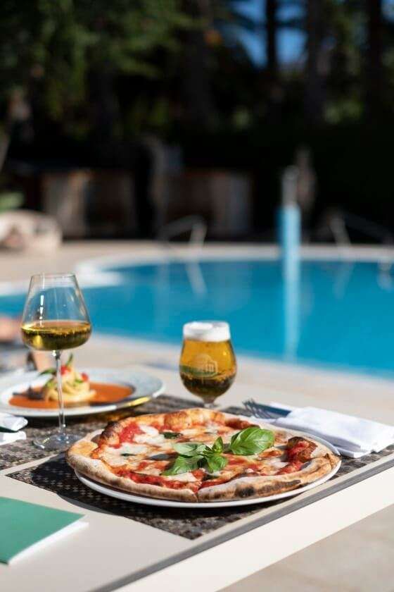 A pizza, a glass of wine, and a glass of beer on a table poolside, with a clear blue swimming pool in the background.