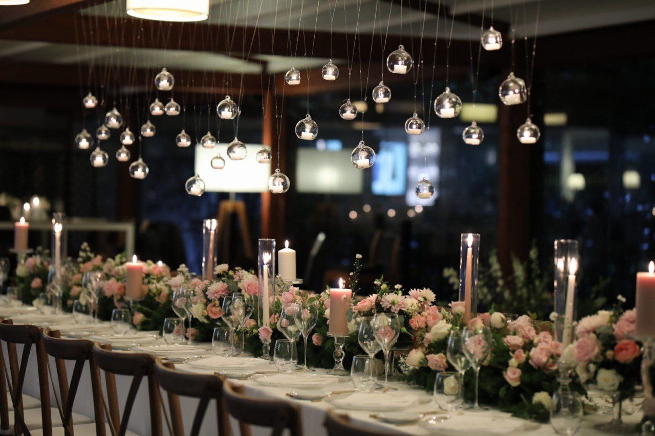 A long banquet table set for a formal dinner with floral centerpieces, candles, and hanging glass globe lights overhead.