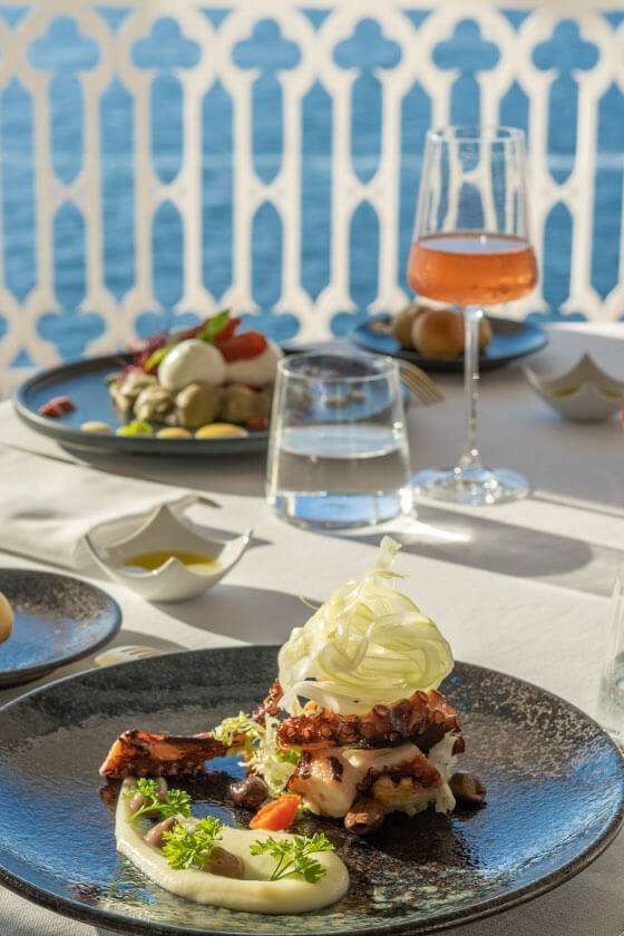 A gourmet meal with octopus and puree sits on a table overlooking the sea, accompanied by a glass of rosé wine.