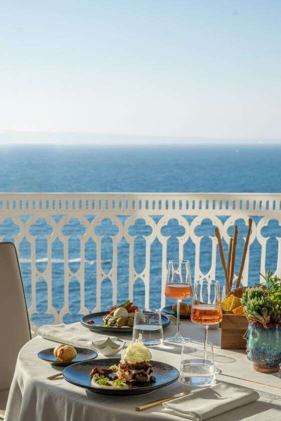 A dining table with two plates of food and glasses of rose wine, set on a balcony overlooking a bright blue sea.