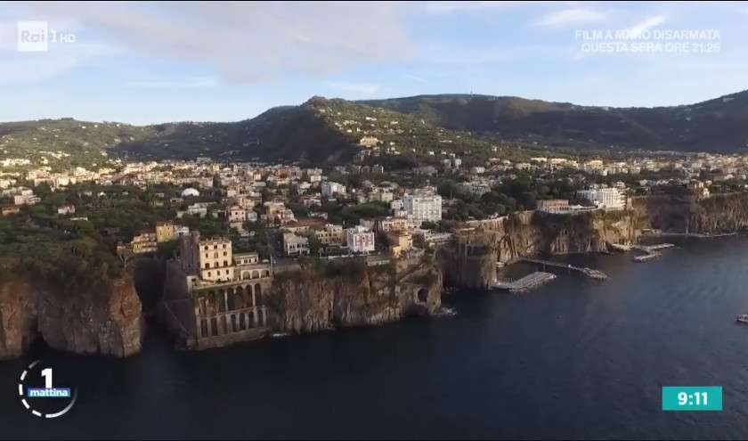 An aerial view of the seaside town of Sorrento, Italy, with buildings perched on cliffs above the dark blue Tyrrhenian Sea.