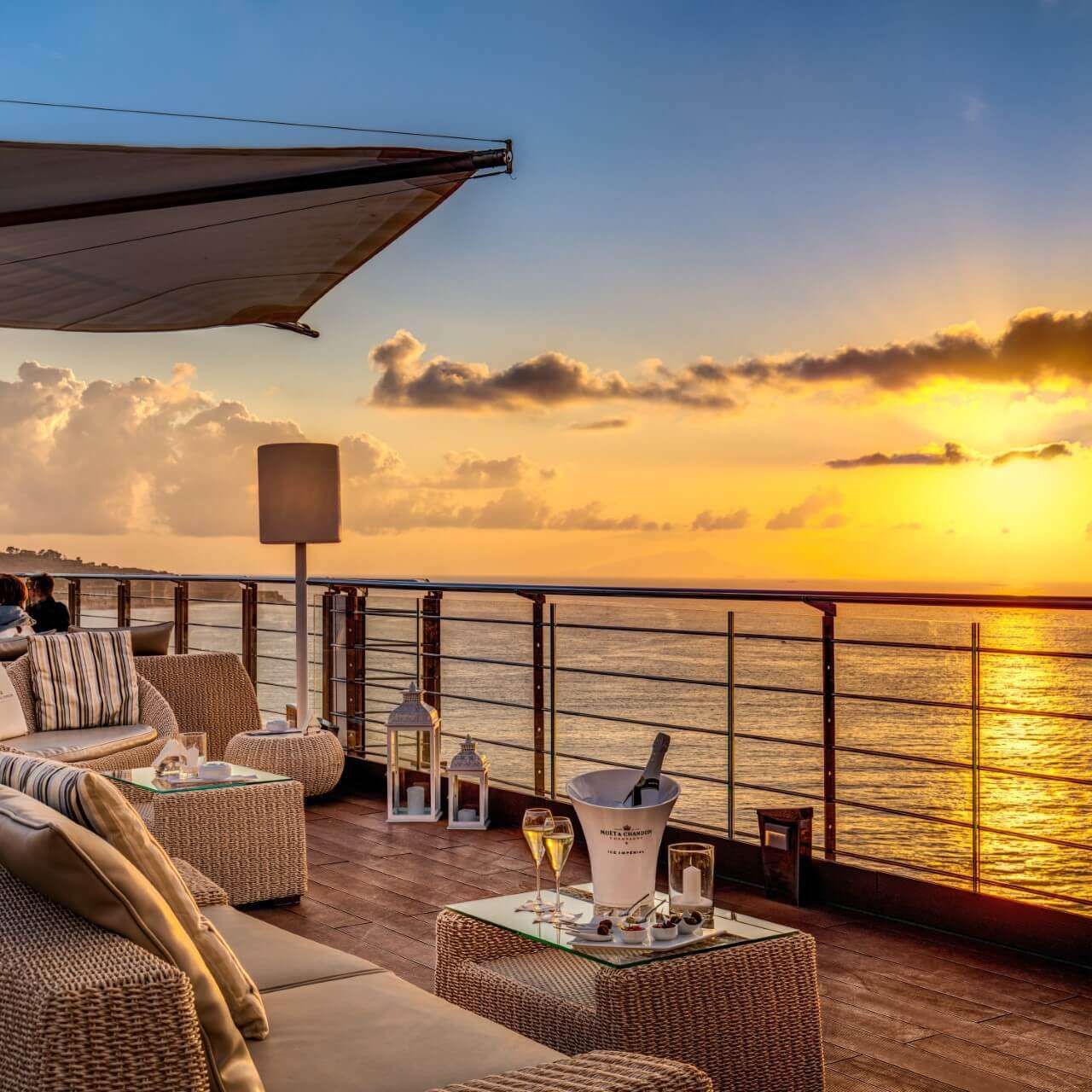 Un patio con balcone affacciato sull'oceano al tramonto, arredato con mobili in vimini, un secchiello per lo champagne e un ombrellone in tessuto.