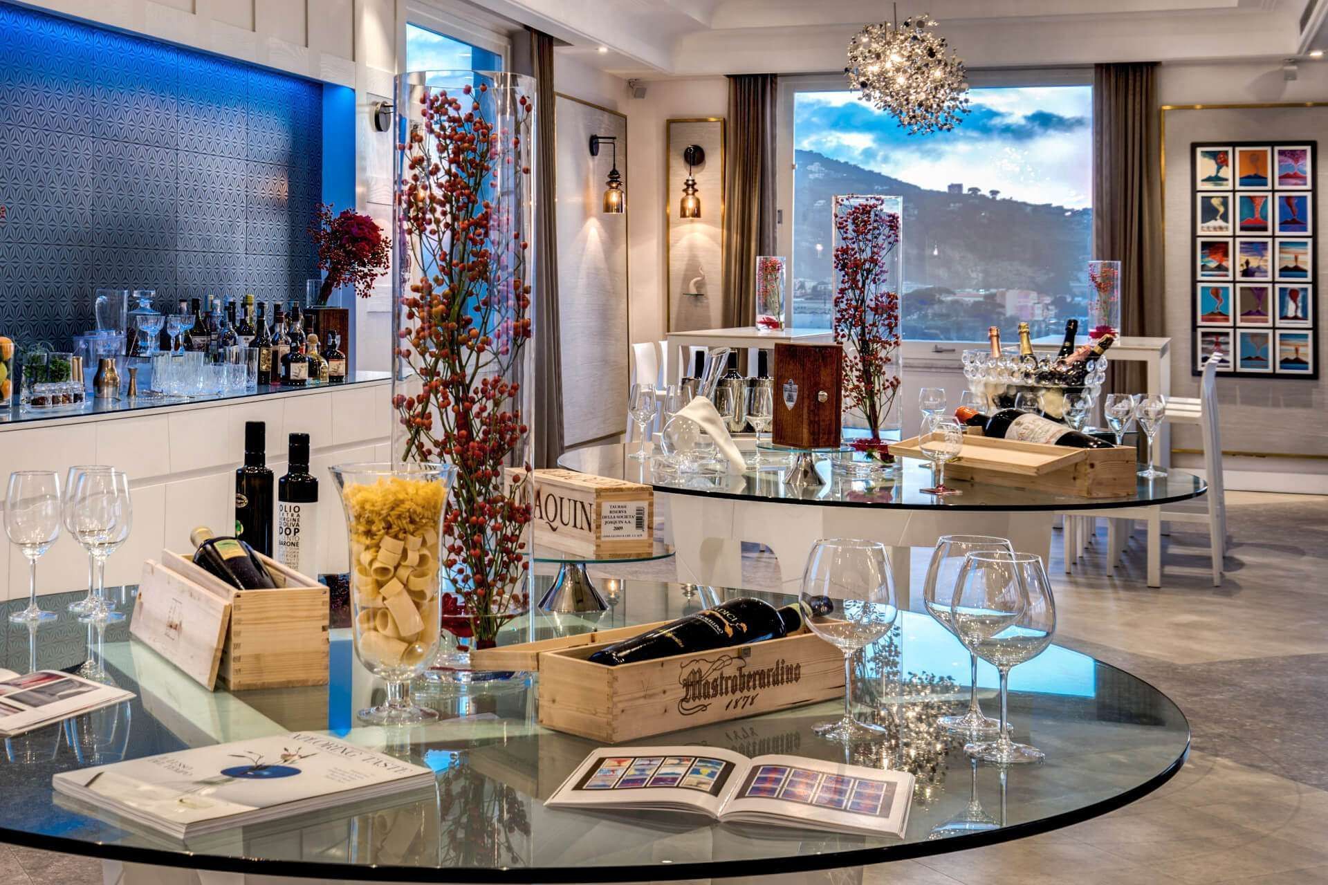 A modern lounge area features glass tables holding wine bottles in wooden crates, glasses, and decor against a harbor view.