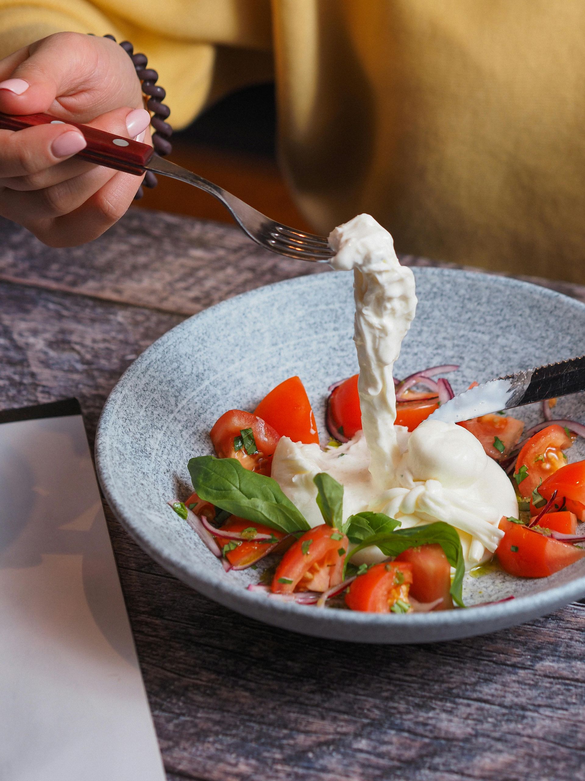 Una mano usa una forchetta per sollevare un pezzo di cremosa burrata da una ciotola di pomodori freschi, basilico e cipolla rossa.