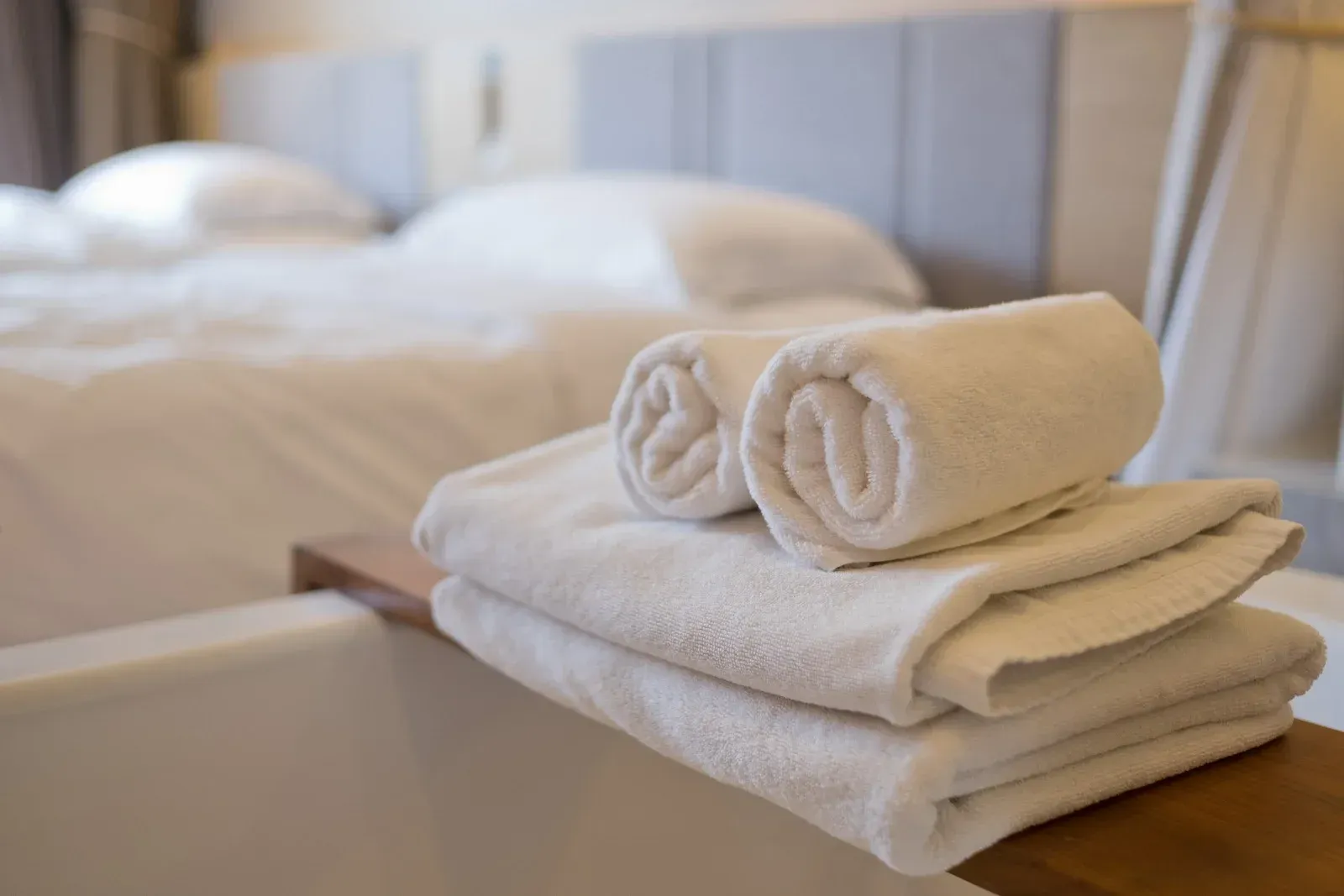 White rolled and folded towels on a wooden tray atop a bathtub, with a bed in the background.