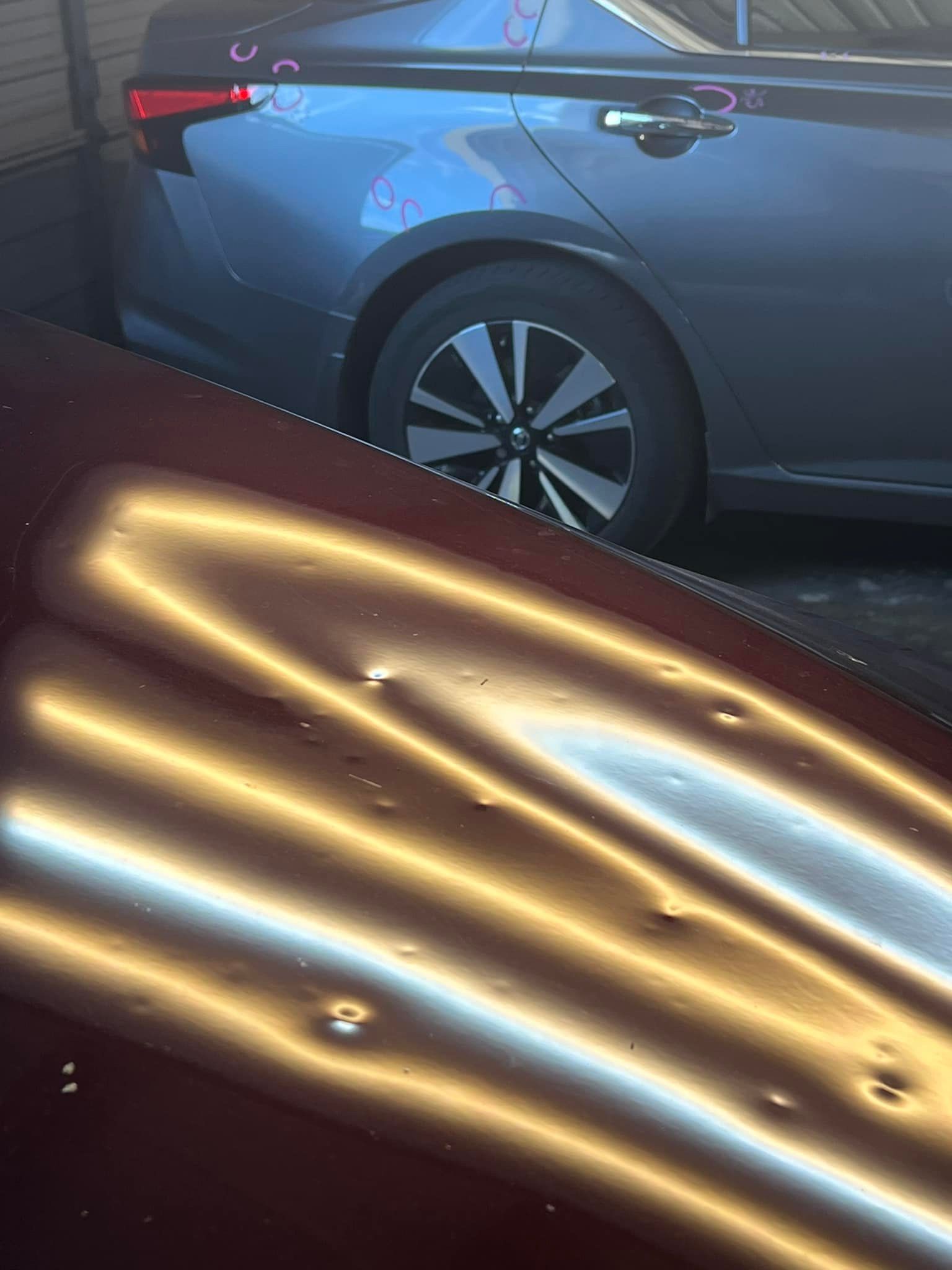 Red car hood with multiple dents, grey car wheel visible in background.