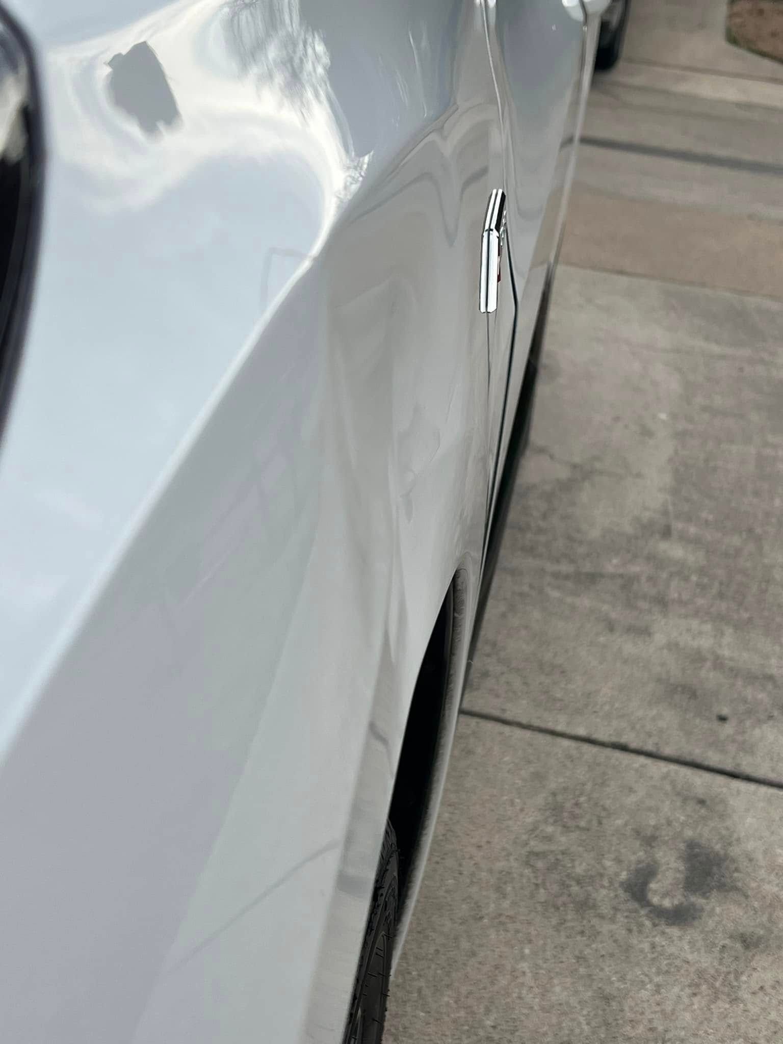 White car's side view with a dent in the door. The car is parked next to a sidewalk.