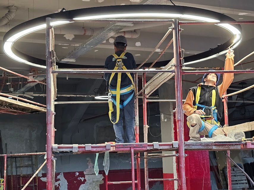 Trabajadores en andamios instalando una gran lámpara de techo circular en una obra de construcción interior.