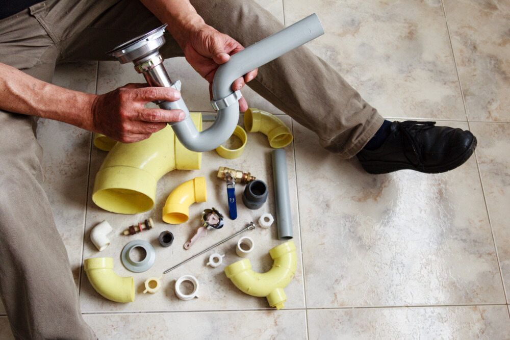 A Man Is Sitting On The Floor Fixing A Sink Pipe — C.J Honeysett Plumbing In Dubbo, NSW