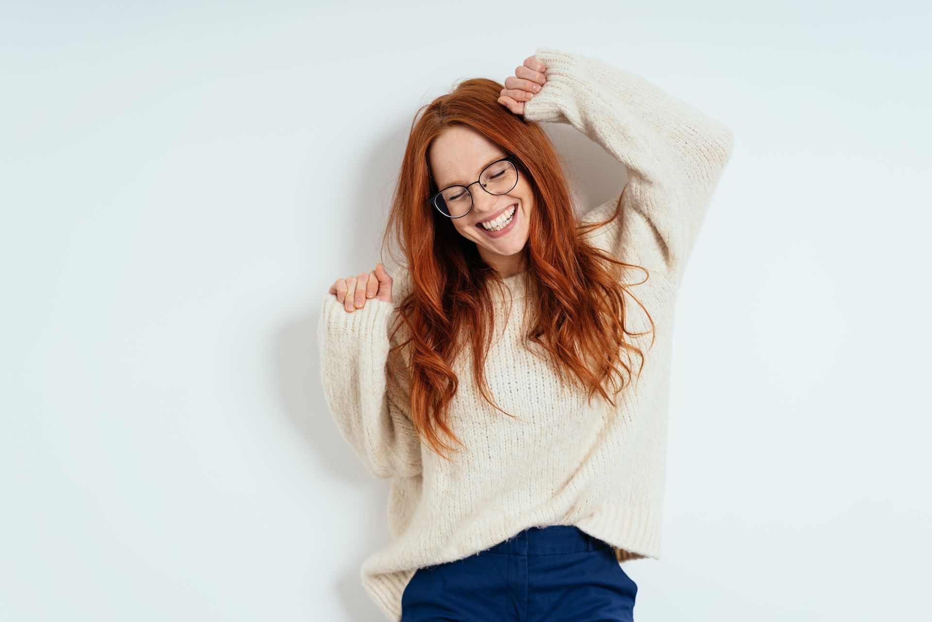 Frau mit roten Haaren, Brille und cremefarbenem Pullover, lächelnd und mit erhobenen Armen, an eine weiße Wand gelehnt.