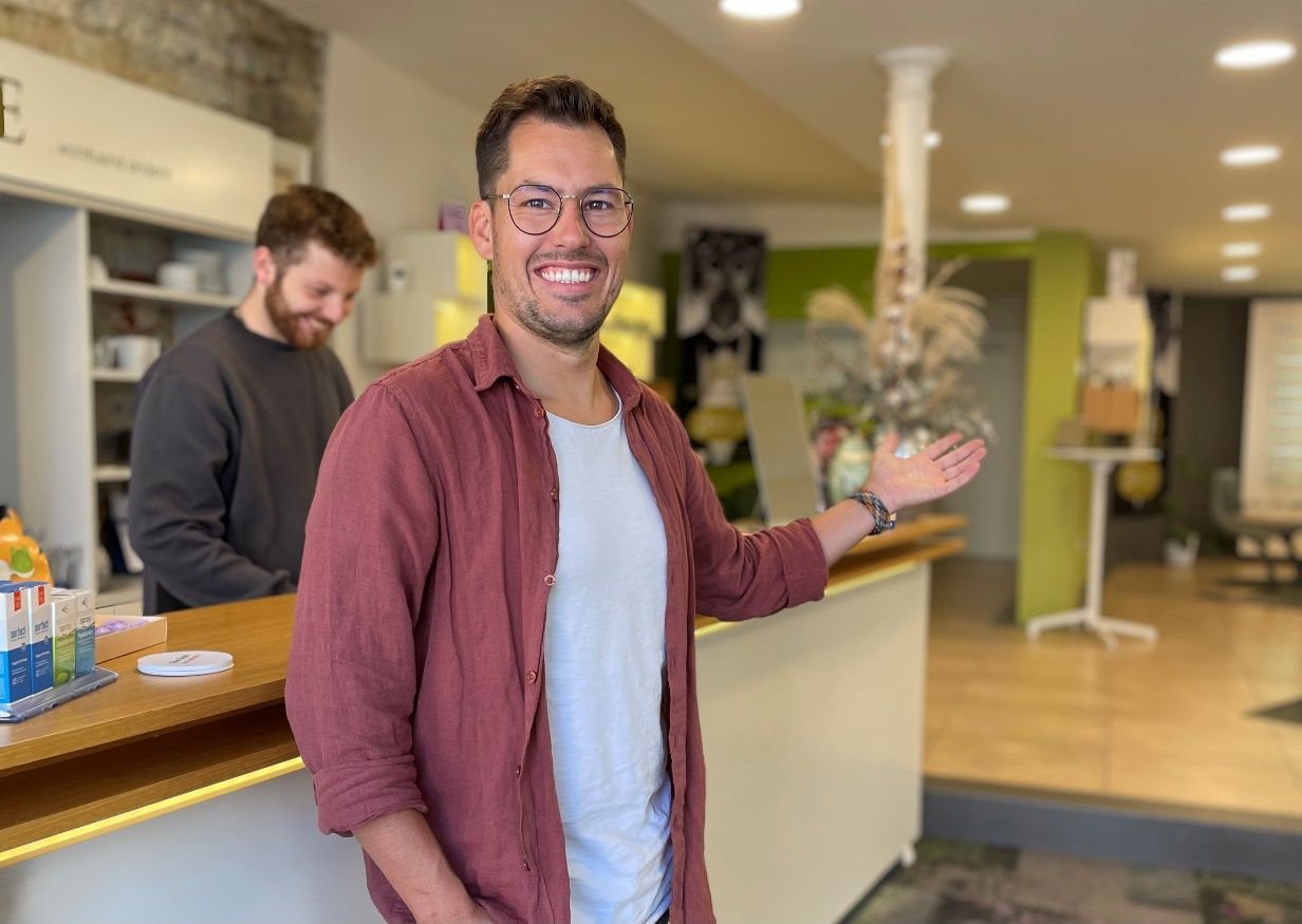 Mann im roten Hemd begrüßt Kunden an einer Ladentheke, ein weiterer Mann im Hintergrund.