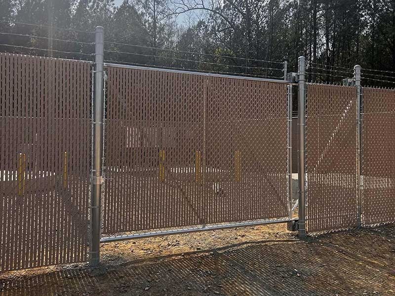 Brown privacy chain-link fence with razor wire at the top, next to trees.