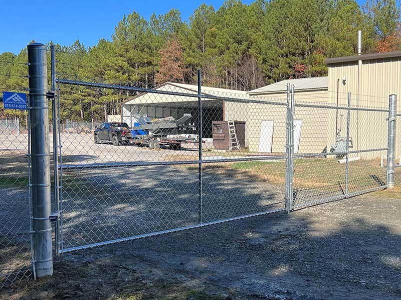 Chain-link fence with a gate open, in front of a building with trees in the background.