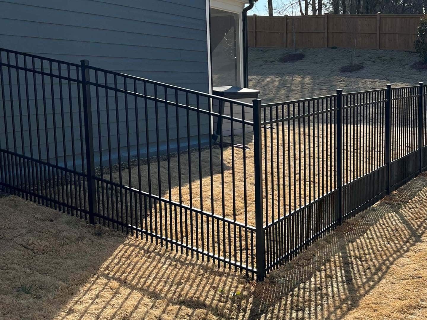 Black metal fence around a yard, casting a shadow on dry grass.