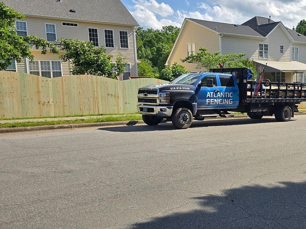Blue Atlantic Fencing truck parked on street in front of a wooden fence, houses in the background.