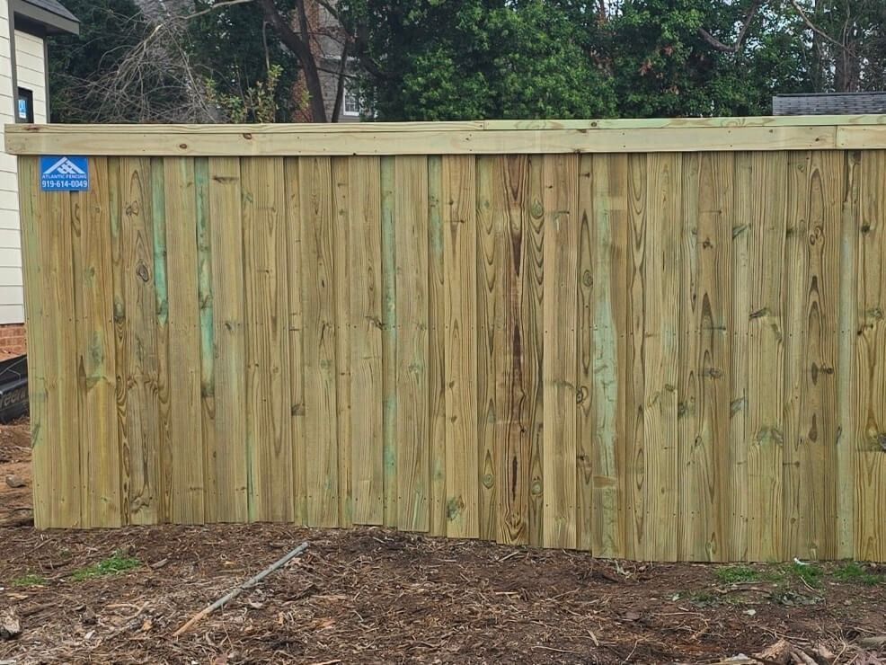 Wooden fence with vertical planks, light green-yellow color, in front of a house and bushes.
