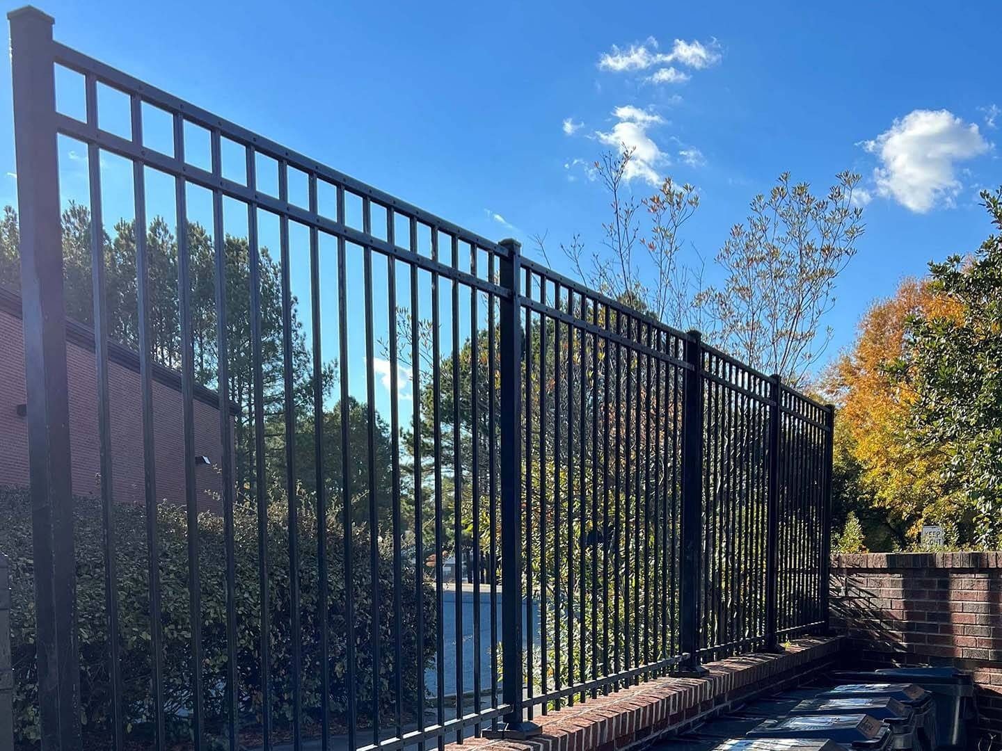 Black metal fence on a brick wall, set against a background of trees and blue sky.