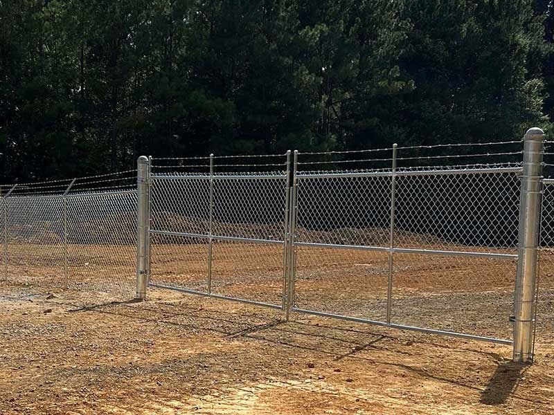 Chain-link fence with barbed wire at top, brown ground, green trees in background.