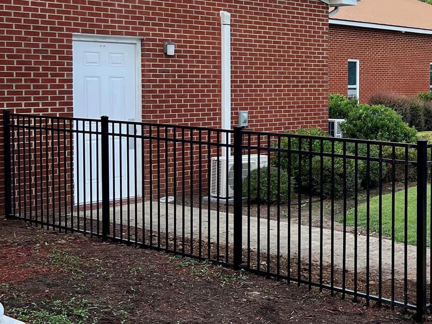 Black metal fence in front of a brick building with a white door and a small hedge.