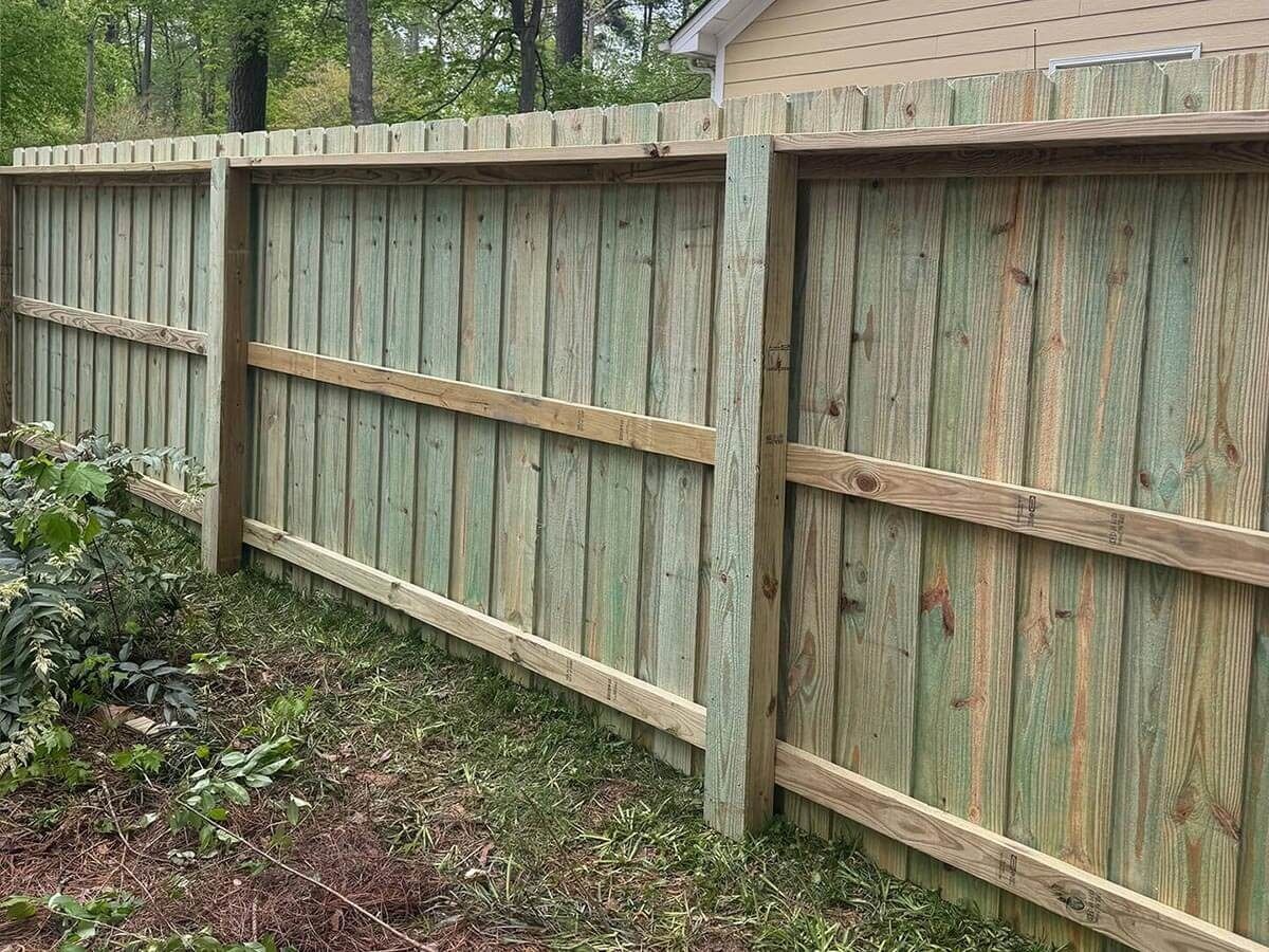 Wooden privacy fence with green-tinted vertical planks and horizontal support beams in a backyard setting.