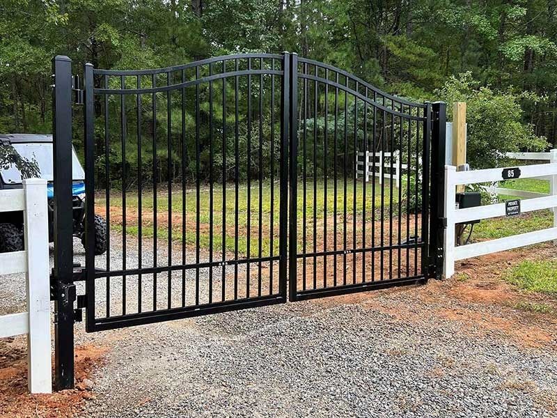 Black wrought-iron double gate set in a gravel driveway, framed by white fences and wooded background.