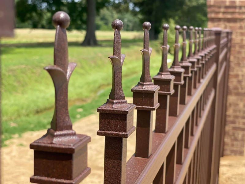 Bronze fence with ornate finials, near brick wall and grassy area.