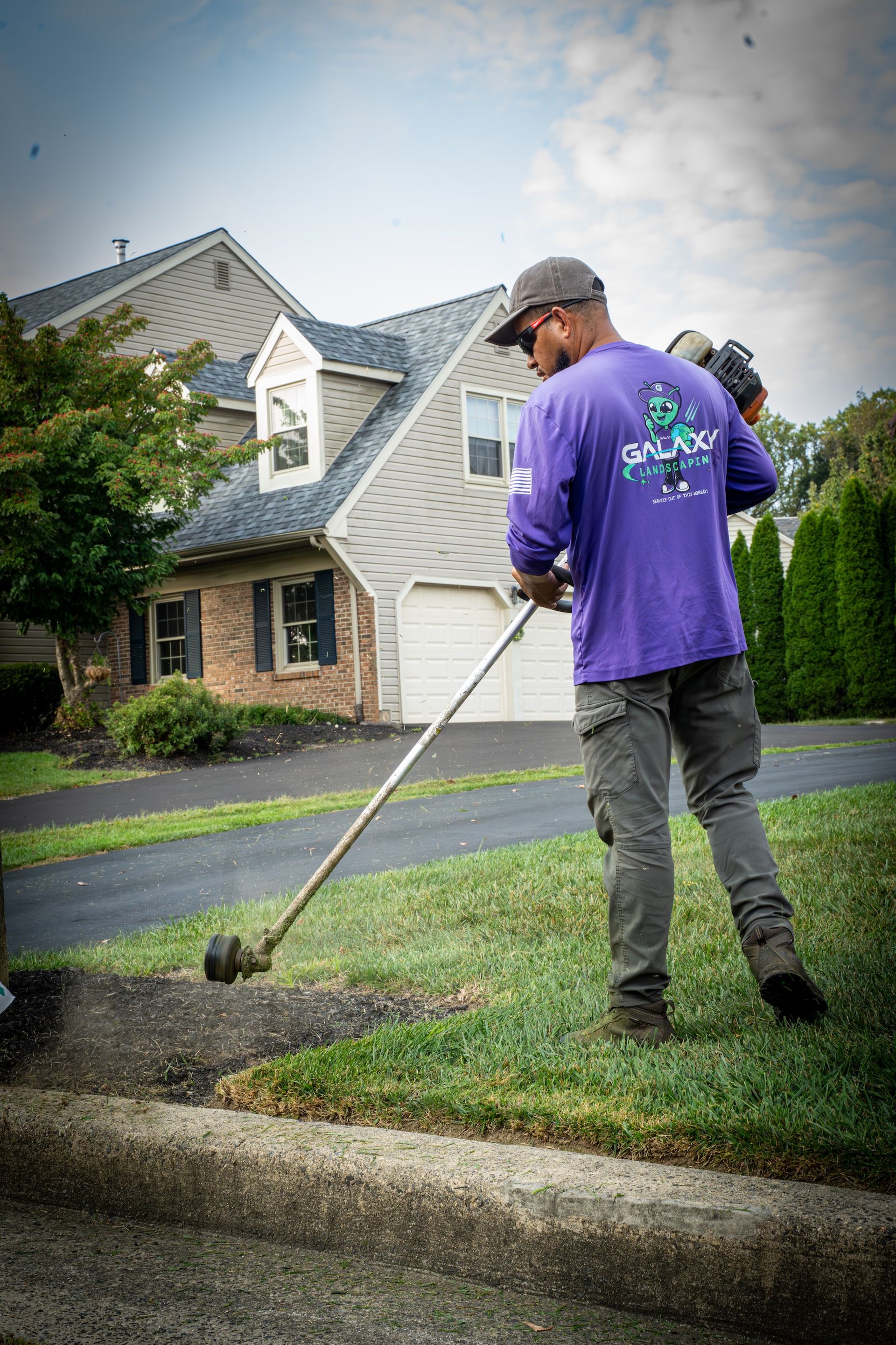 Man in purple shirt trimming grass with a weed whacker near a driveway and house.