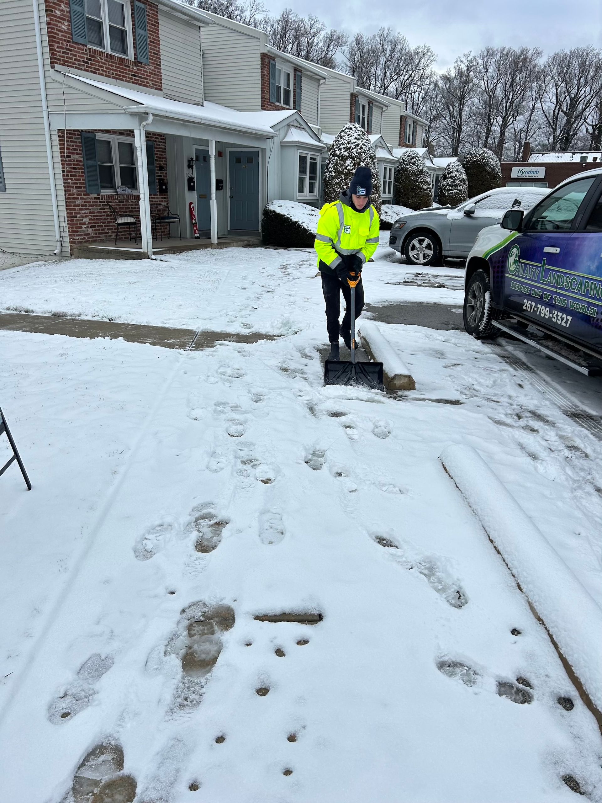 Person in a neon vest shoveling snow from a driveway in front of townhouses. Cars and snow-covered ground are visible.
