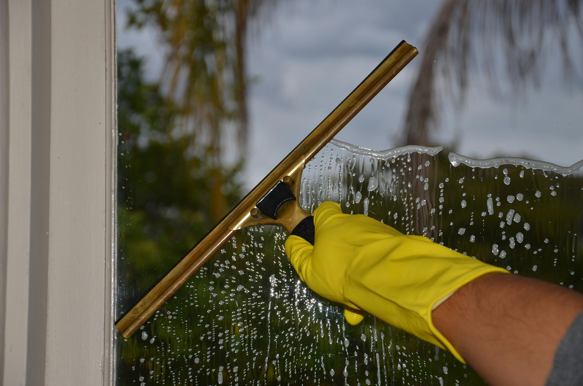 A hand in a yellow rubber glove uses a squeegee to clean soap suds off a window pane.
