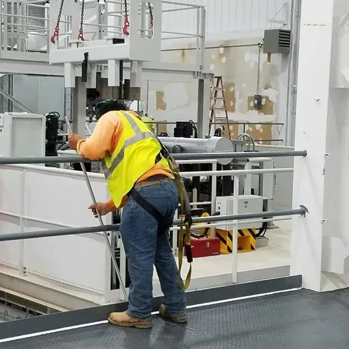 Worker in orange vest leaning on rail inside an industrial facility, inspecting machinery.