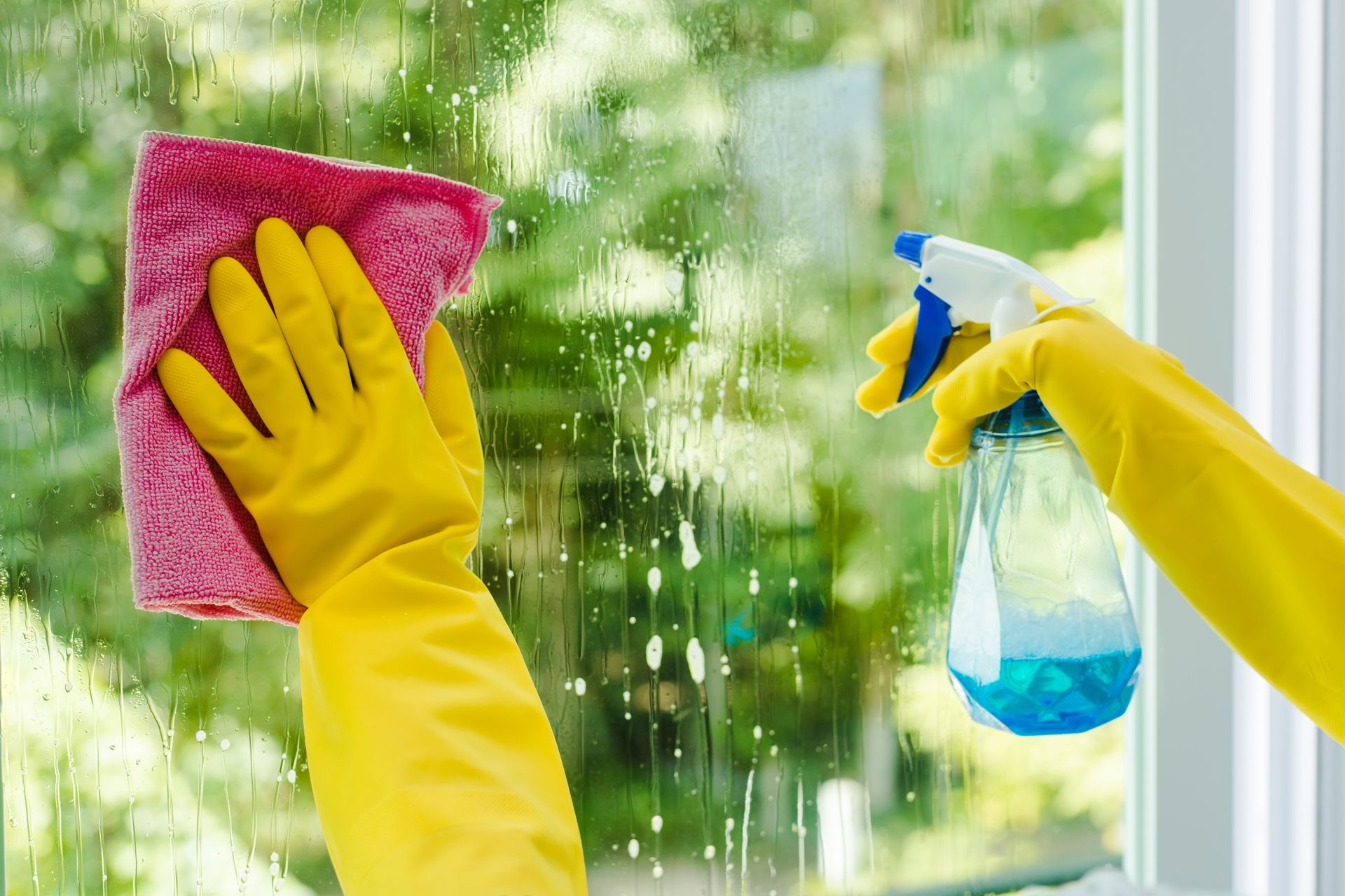 A person wearing yellow gloves is cleaning a window with a cloth and spray bottle.