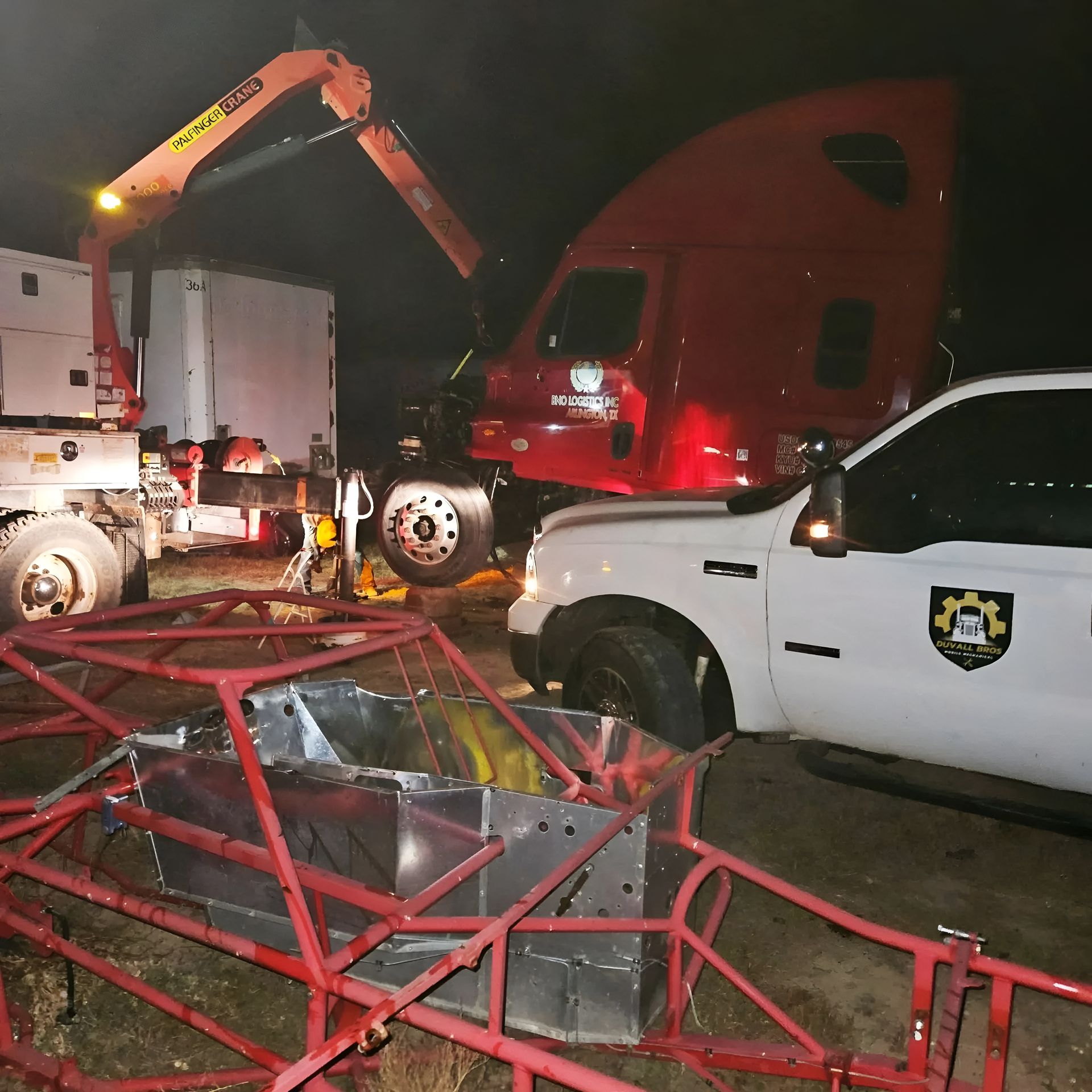 Nighttime crash scene with tow truck, red semi, white police truck, and overturned red metal structure