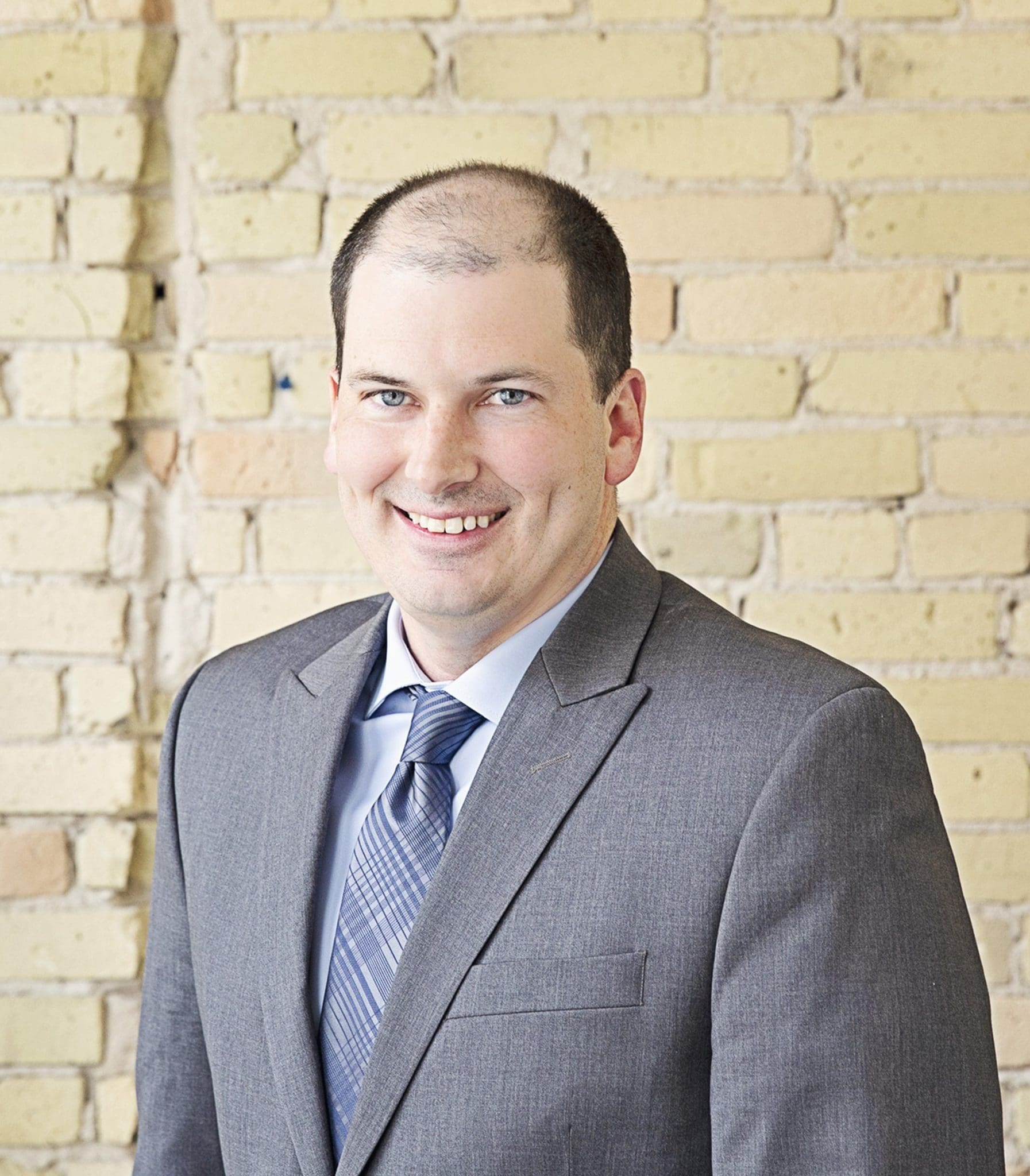 Man in gray suit and patterned tie smiling in front of a brick wall.
