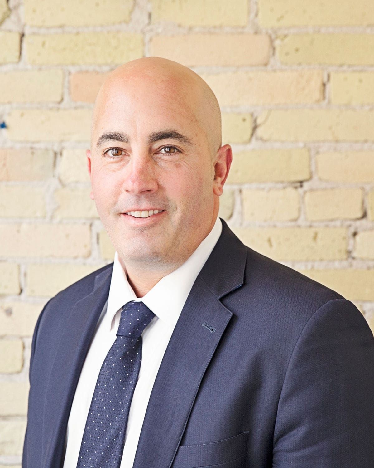 Bald man in a suit smiles, posing in front of a brick wall.