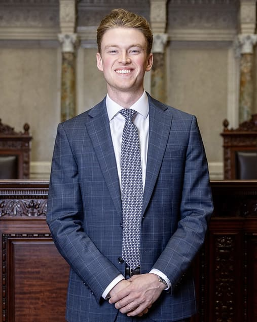 Man in suit smiling, arms crossed. Standing in a wood-paneled room with columns.