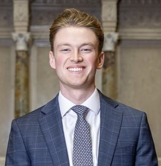 Smiling man in a blue plaid suit and patterned tie, indoors with columns in the background.