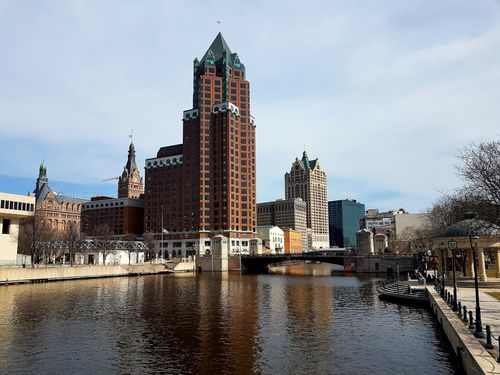 Milwaukee skyline with brown skyscraper and reflections in the river, under a blue sky.