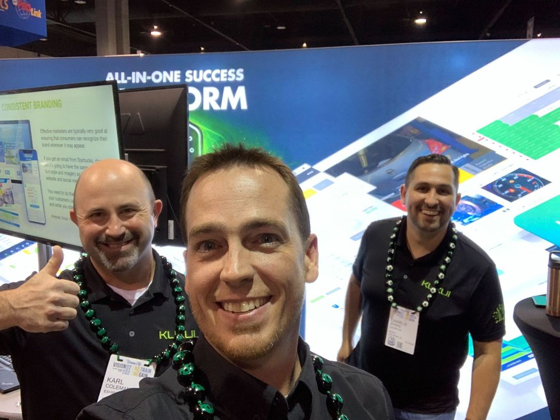 Three smiling men at a trade show booth, with a blue “All-in-one success platform” backdrop.