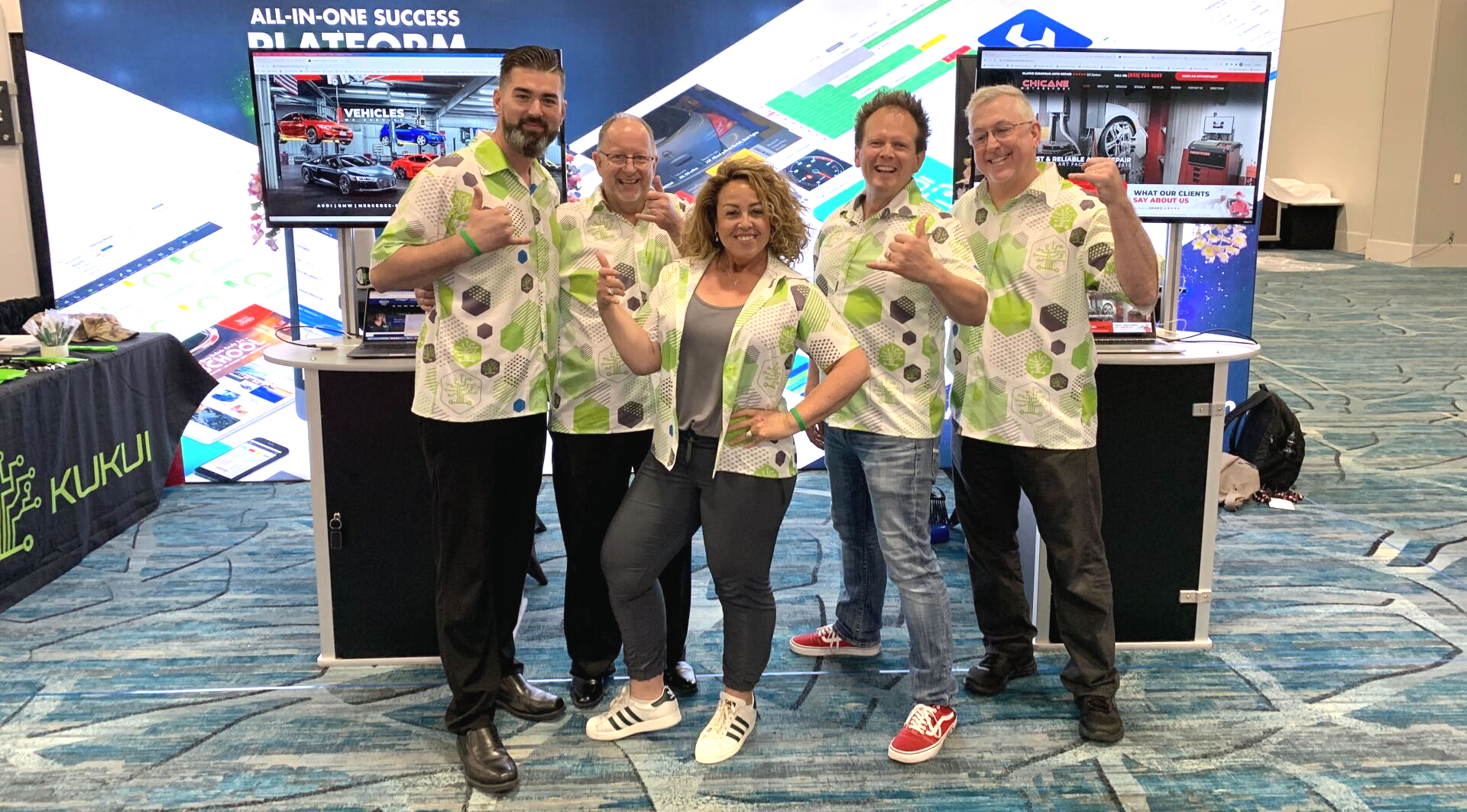 Five people posing at a trade show booth, smiling and holding drinks, with bright displays behind them.