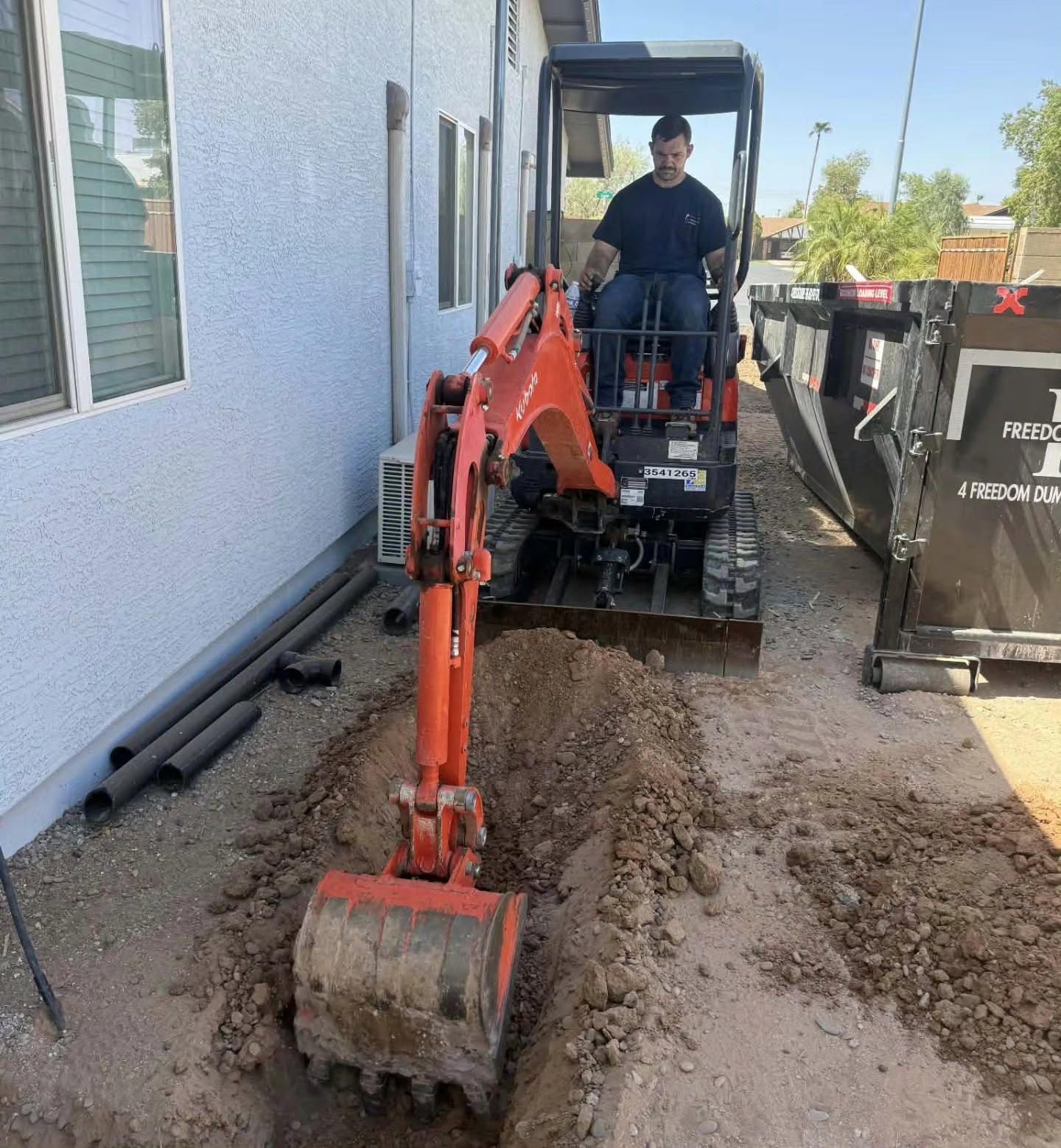 Mini excavator digging a trench beside a house, with a worker operating it and a dumpster nearby.