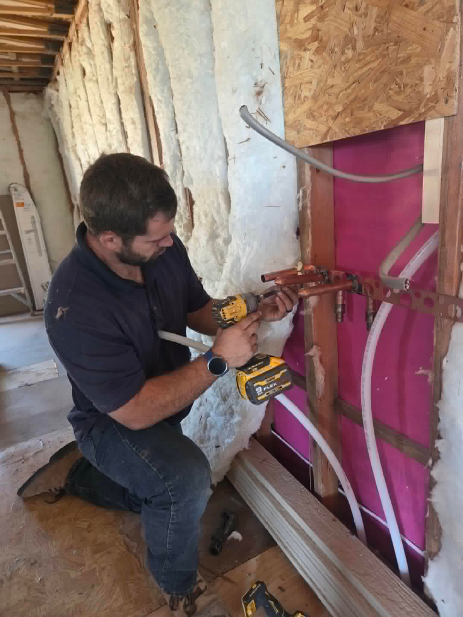 Worker kneeling, using a drill to install copper plumbing in an unfinished wall with pink insulation.