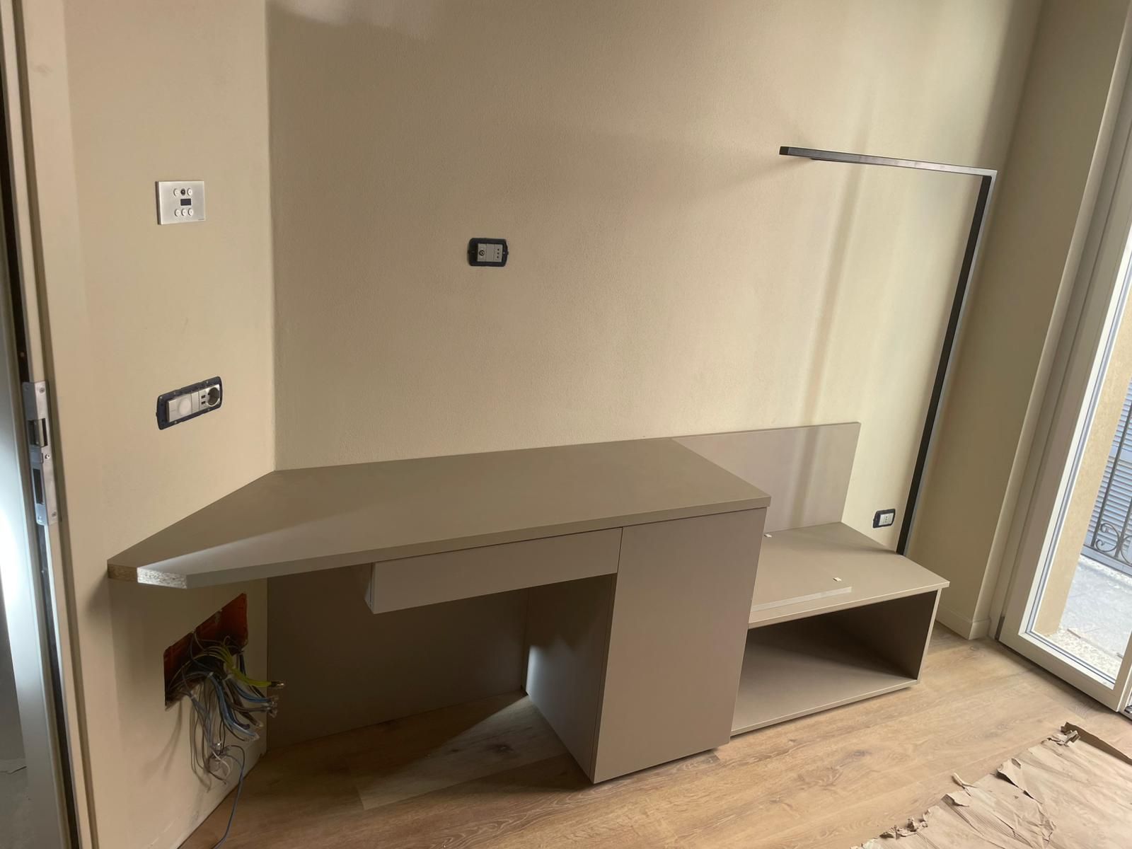 Scrivania a muro beige integrata con ripiani, situata in una stanza vicino a una finestra e a prese elettriche.