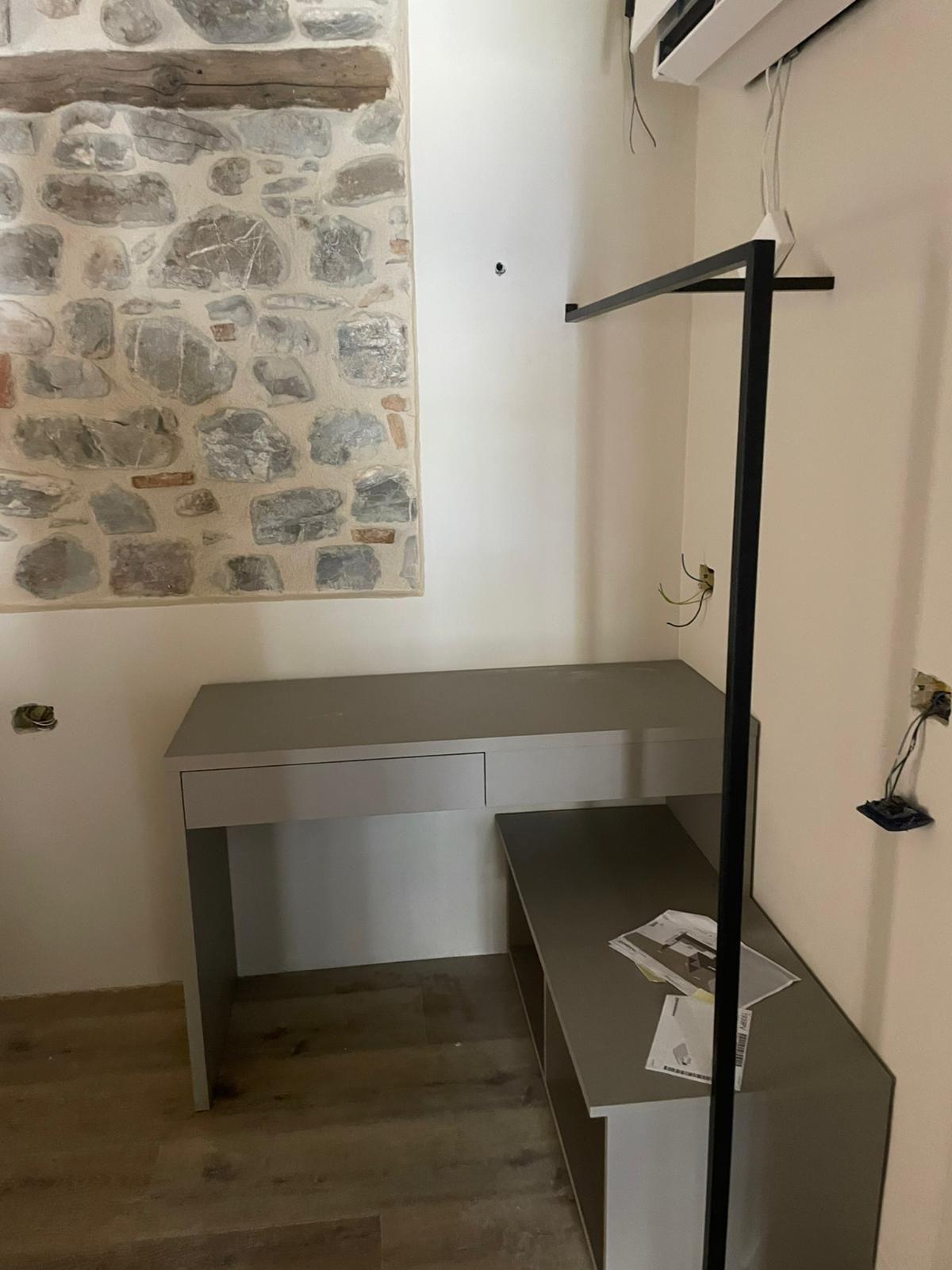 Piccola stanza vuota con parete in pietra, scrivania grigia e scaffale nero su pavimento in legno.