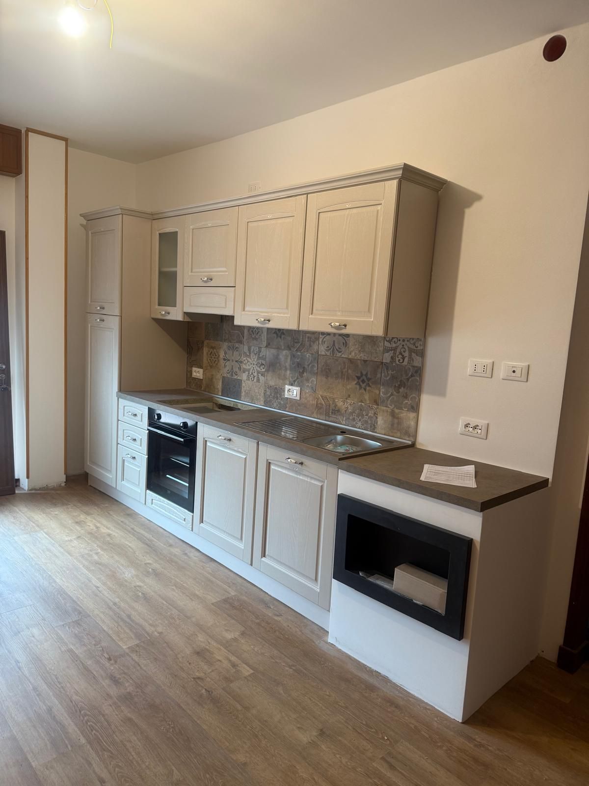 Piccola cucina moderna con mobili beige, paraschizzi piastrellato, pavimento in legno e forno e microonde a incasso.