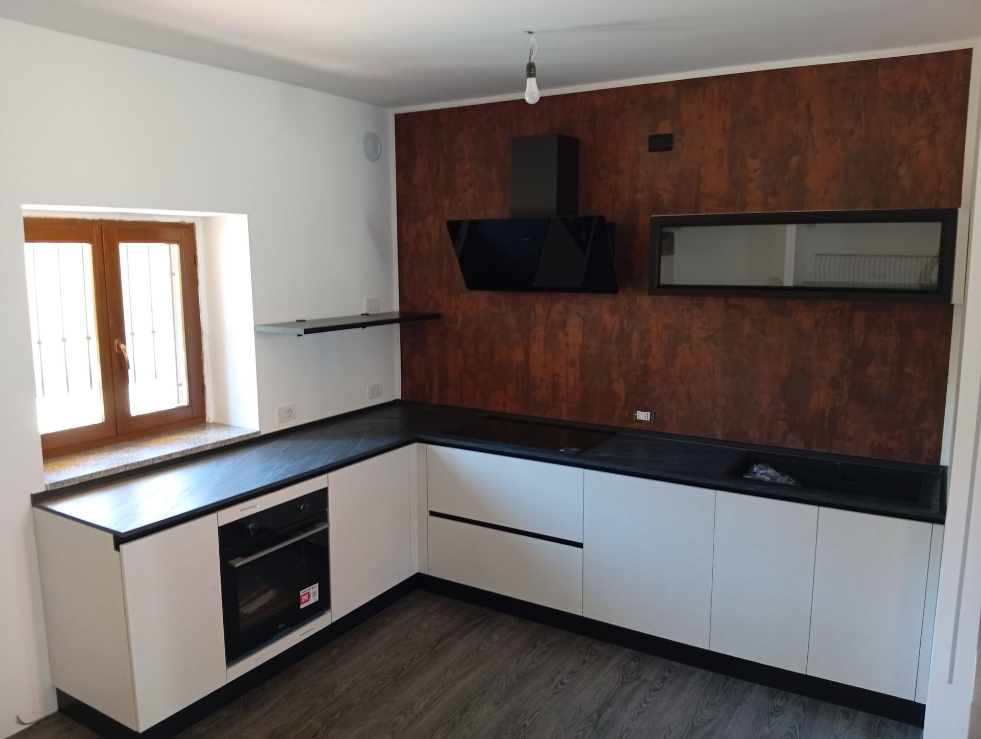 Cucina moderna vuota con mobili bianchi, ripiani neri, parete decorativa in legno e finestra sulla sinistra.