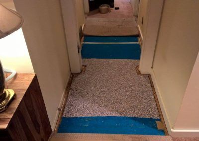 Hallway with carpet partially removed, exposing blue and tan flooring. Walls are white.