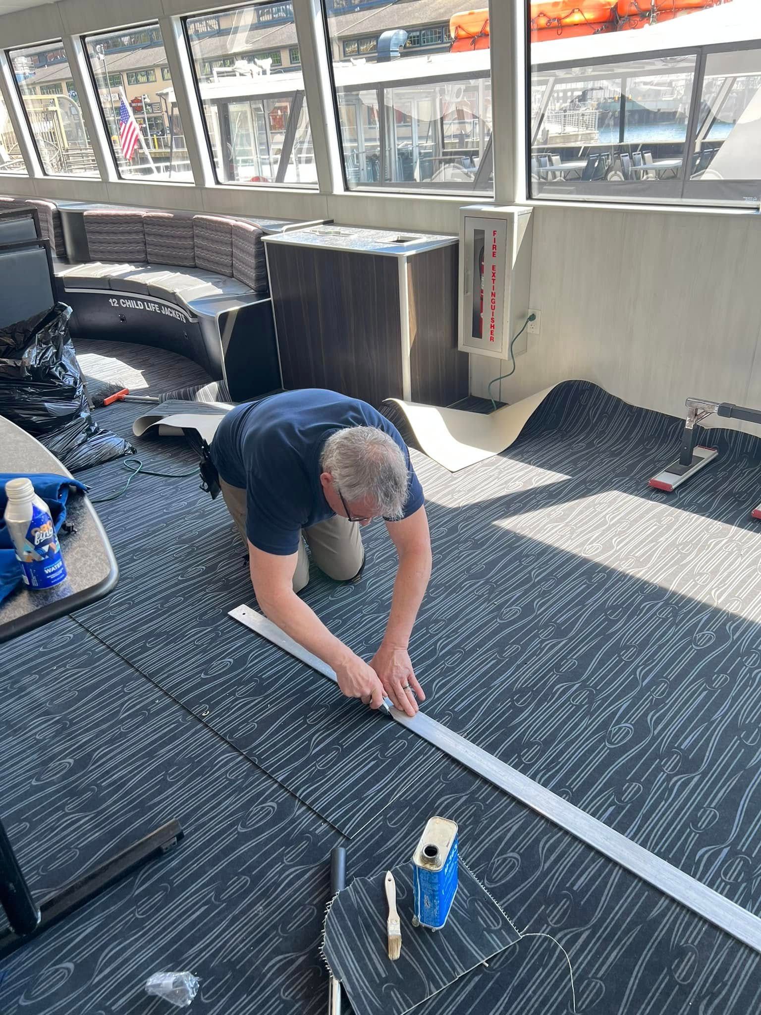 Man kneeling, cutting carpet with a utility knife inside a room with large windows; rolls of carpet and tools are visible.