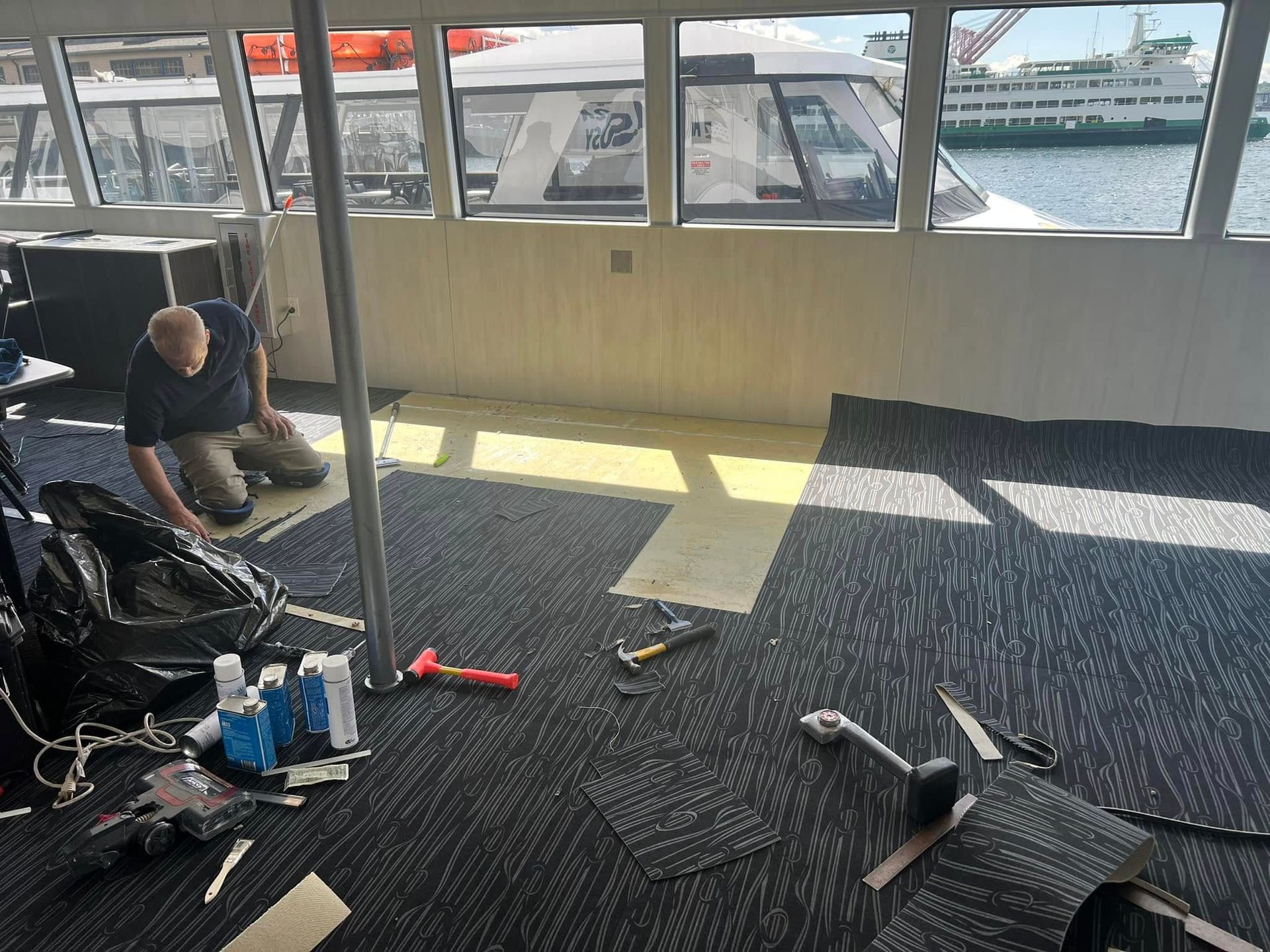 Person installing carpet tiles inside a ferry. Tools and materials scattered around. Sunny day, harbor view.